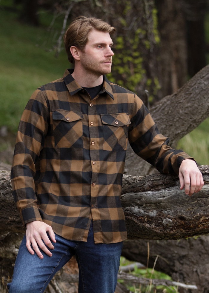 A man wearing a black and brown buffalo check flannel shirt with two flap breast pockets and two button cuff, standing next to a fallen tree.