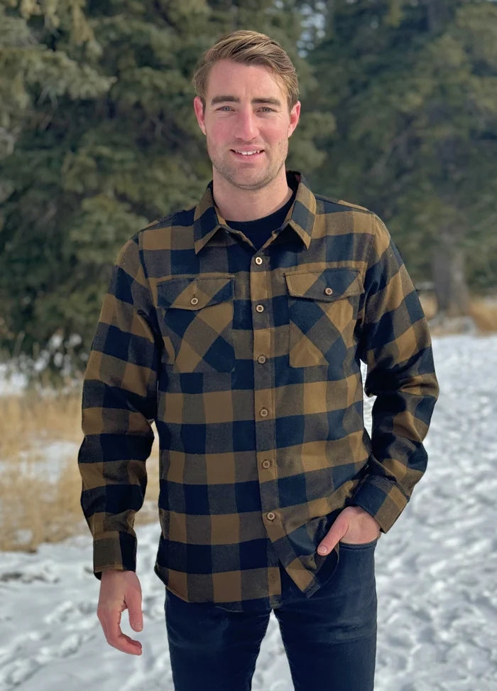 A man wearing a black and brown buffalo check flannel shirt with two flap breast pockets and two button cuff, standing with one hand in his front pocket.