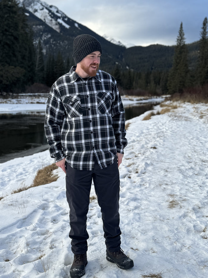 A man wearing a black and white plaid padded flannel jacket, standing near a snowy landscape.