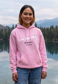 Person wearing a pink hoodie with 'Banff Canada' design by a lake and mountains.