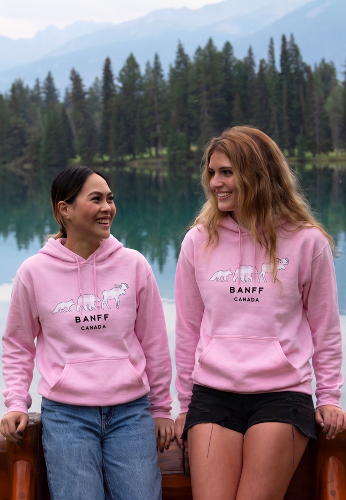 Two people wearing pink Banff hoodies by a lake with trees in the background