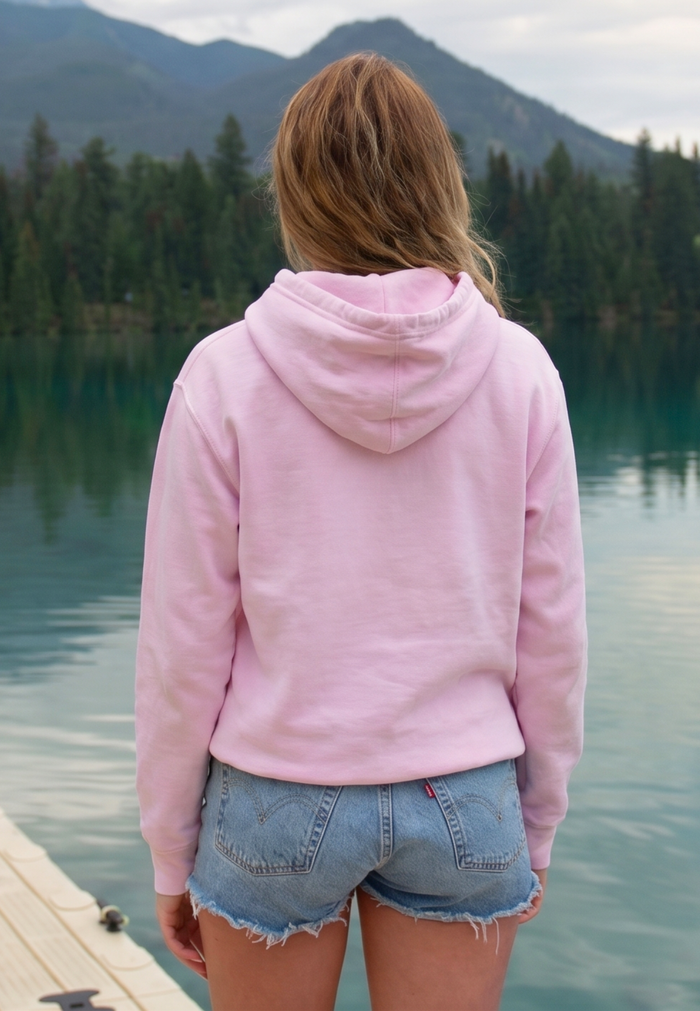 Person wearing a pink hoodie and denim shorts standing by a lake with mountains in the background