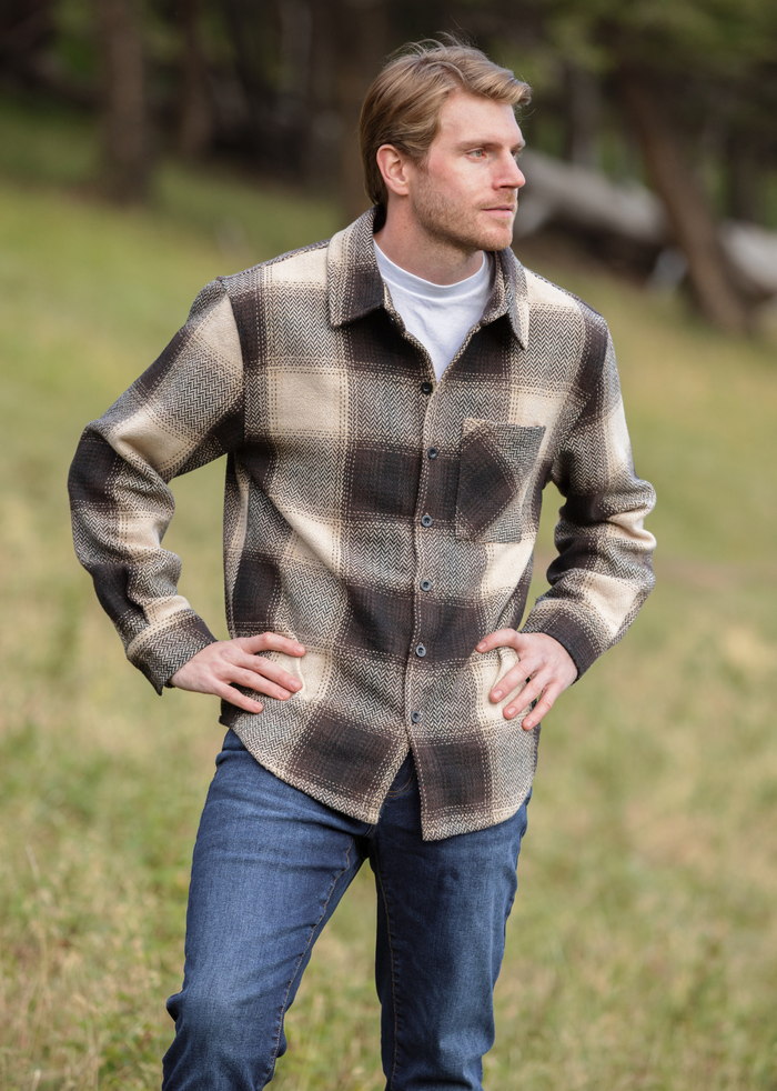 A person wearing a brown wool blend flannel jacket with herringbone weave, paired with blue jeans, standing in a natural outdoor setting.