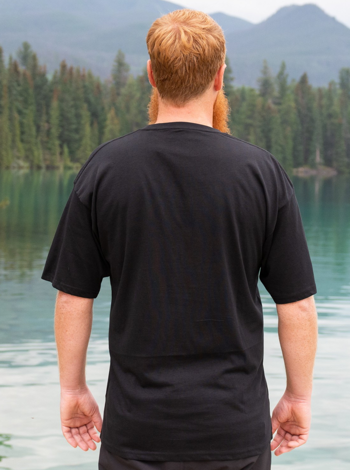 Man wearing a black t-shirt standing by a lake with mountains in the background