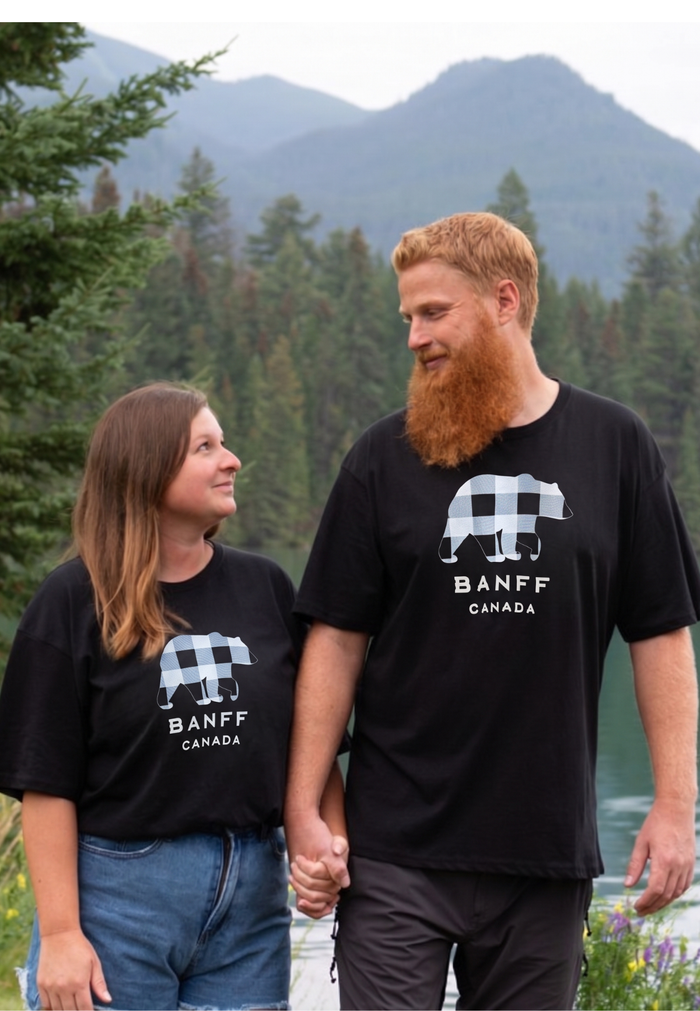 Two people wearing black Banff t-shirts standing in a forested area with mountains in the background.