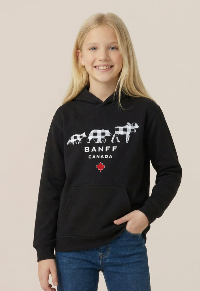 A young girl with blonde hair smiling while wearing a black pullover hoodie with a white Banff Canada graphic and blue jeans, set against a solid light beige background.