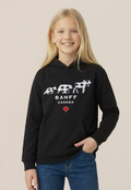 A young girl with blonde hair smiling while wearing a black pullover hoodie with a white Banff Canada graphic and blue jeans, set against a solid light beige background.