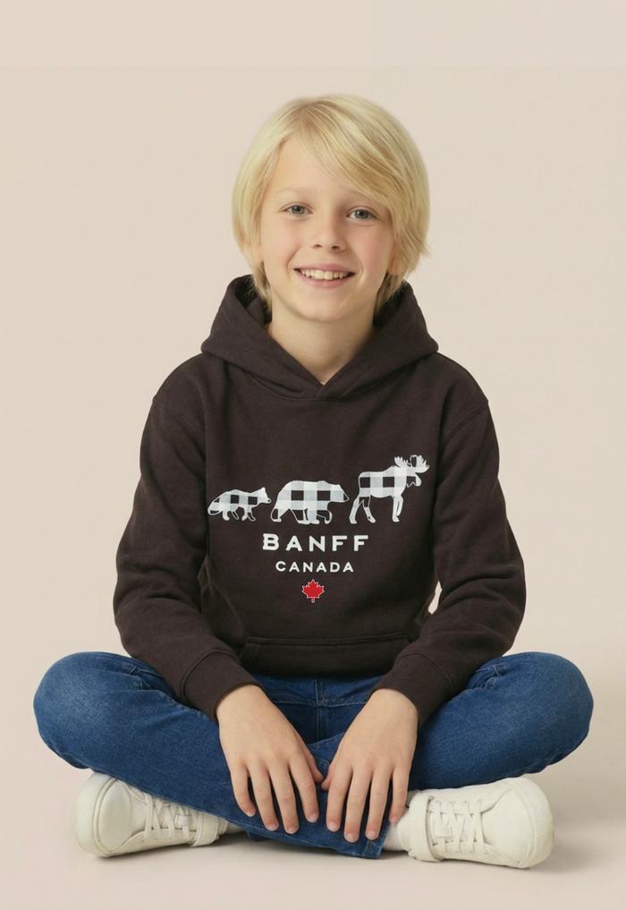 A young blonde boy sitting cross-legged wearing a dark brown Banff Canada hoodie, blue jeans, and white sneakers against a solid beige background.