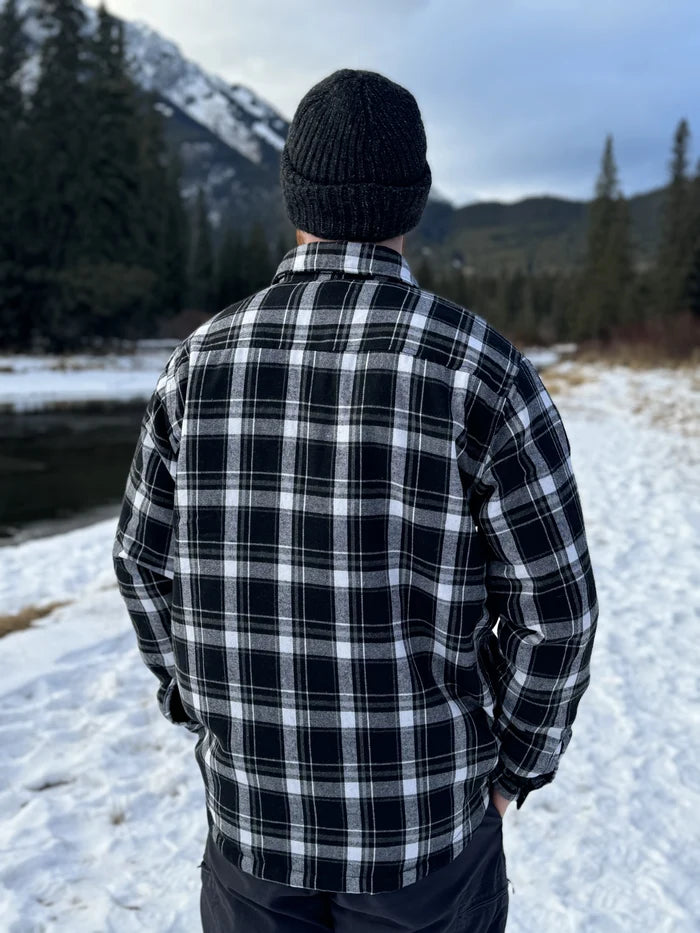 Back view of a man wearing a black and white plaid padded flannel jacket, standing near a snowy landscape.