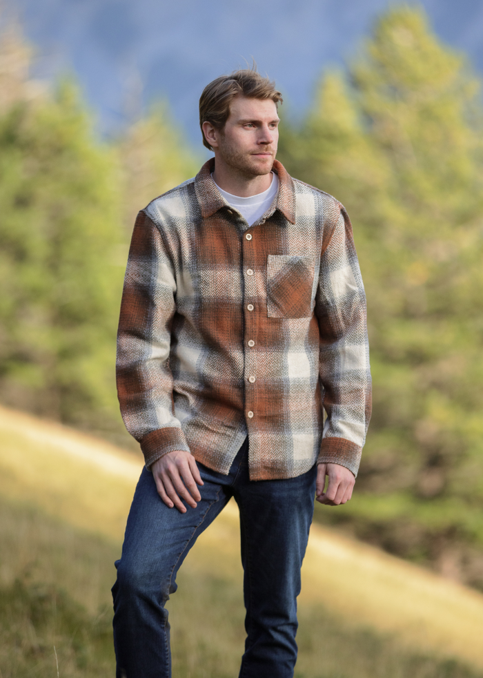 A person standing outdoors wearing a plaid flannel shacket with a herringbone weave in a blend of brown and blue colors.