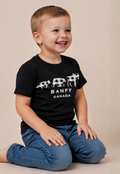 A young boy smiling and looking to the side, wearing a black Banff Canada t-shirt with a white buffalo graphic and blue jeans, sitting against a plain beige background.