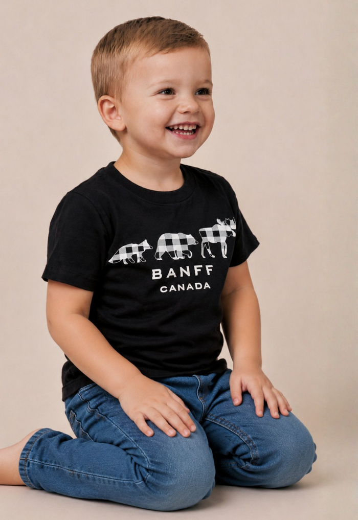 A young boy smiling and looking to the side, wearing a black Banff Canada t-shirt with a white buffalo graphic and blue jeans, sitting against a plain beige background.