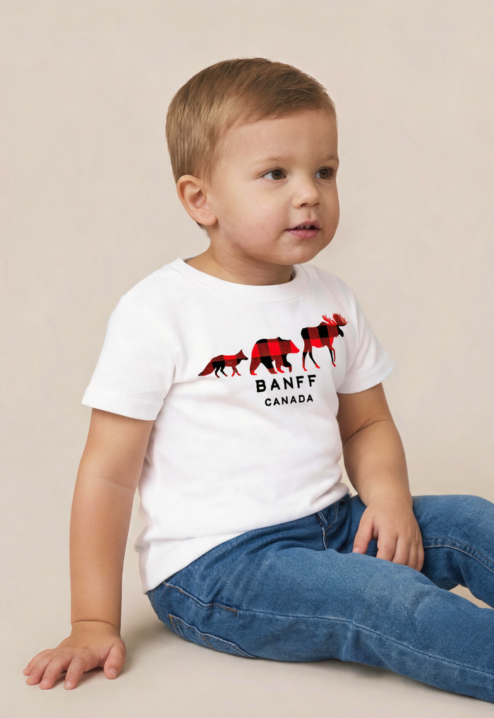 A toddler boy wearing a white short-sleeve t-shirt with a red and black moose graphic and the text 'BANFF CANADA', paired with blue jeans, sitting against a plain beige background.
